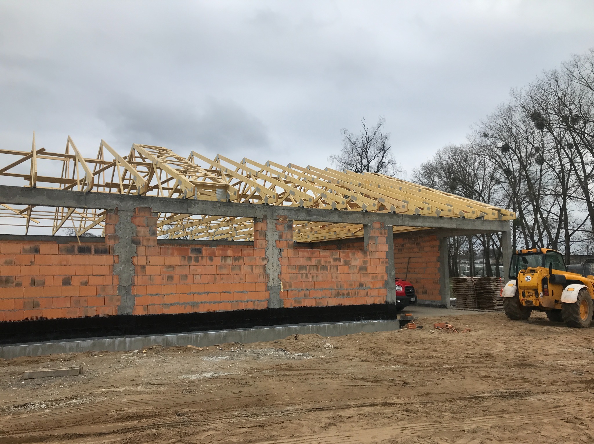 Budowa domu jednorodzinnego w stanie surowym otwartym: ściany z czerwonej cegły, konstrukcja dachu z drewna, żółta koparko-ładowarka na placu budowy.