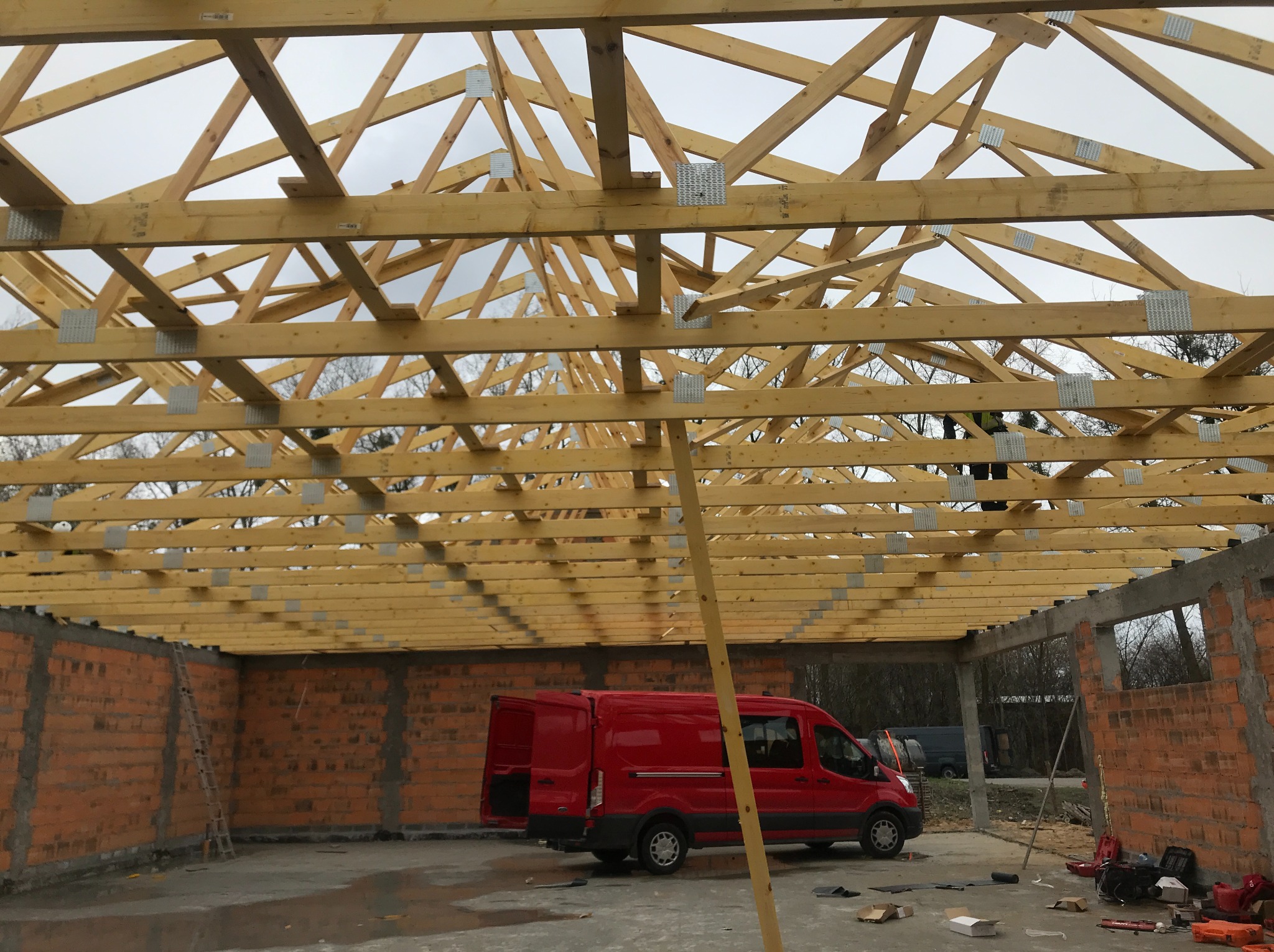 Wnętrze budynku w trakcie budowy. Widoczna drewniana konstrukcja dachu z metalowymi wzmocnieniami. Czerwony van zaparkowany wewnątrz, ceglane ściany w tle.