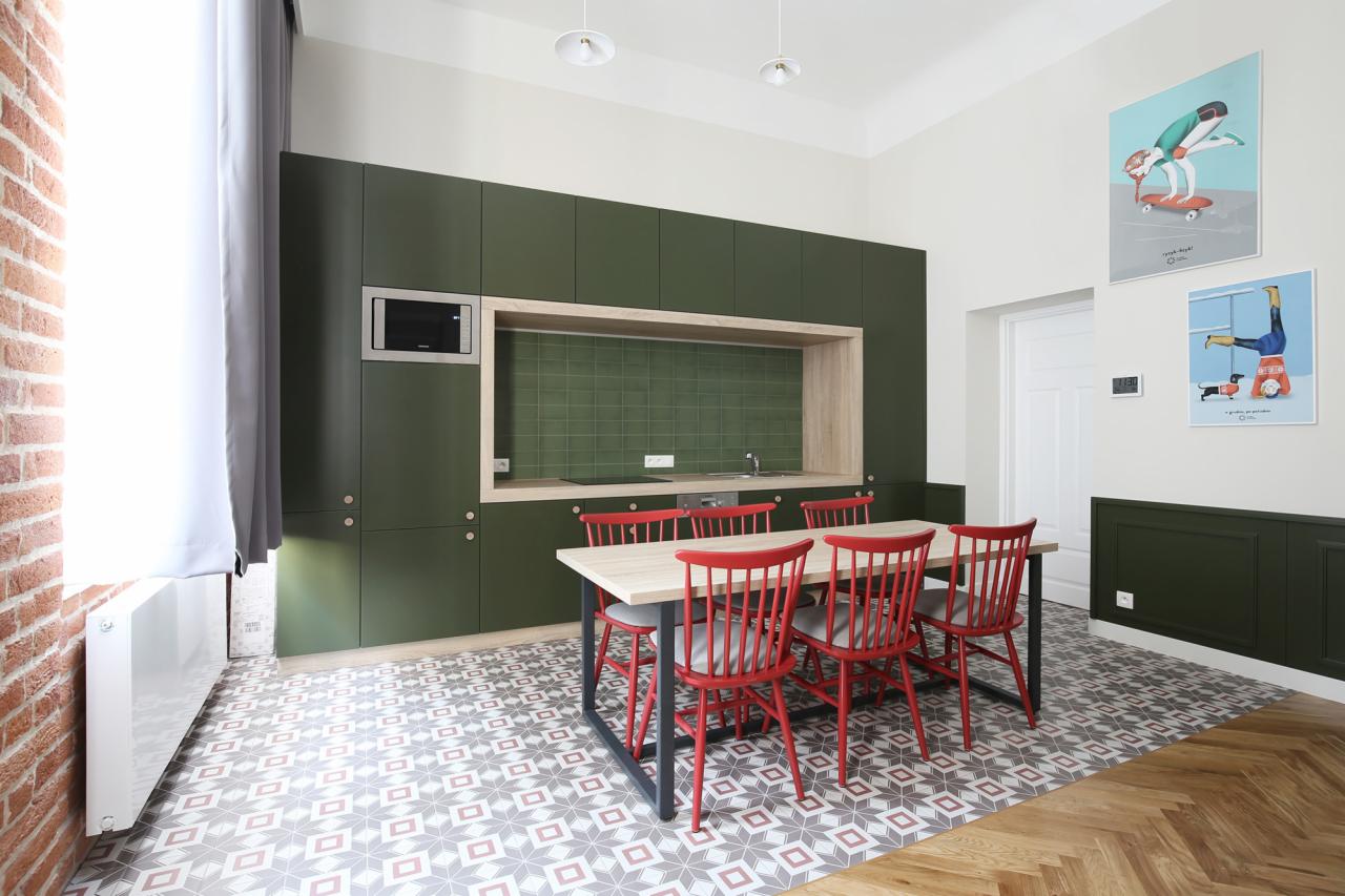 Nowoczesna kuchnia z ciemnozielonymi szafkami, drewnianym blatem i czerwonymi krzesłami, na ścianie grafiki.