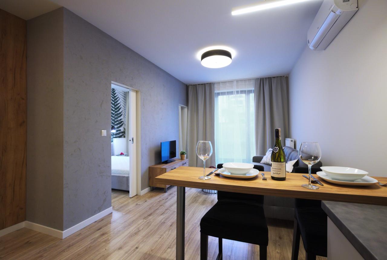 Nowoczesne wnętrze apartamentu z drewnianą podłogą, stołem nakrytym dla dwóch osób, butelką wina i widokiem na sypialnię z łóżkiem oraz telewizor.