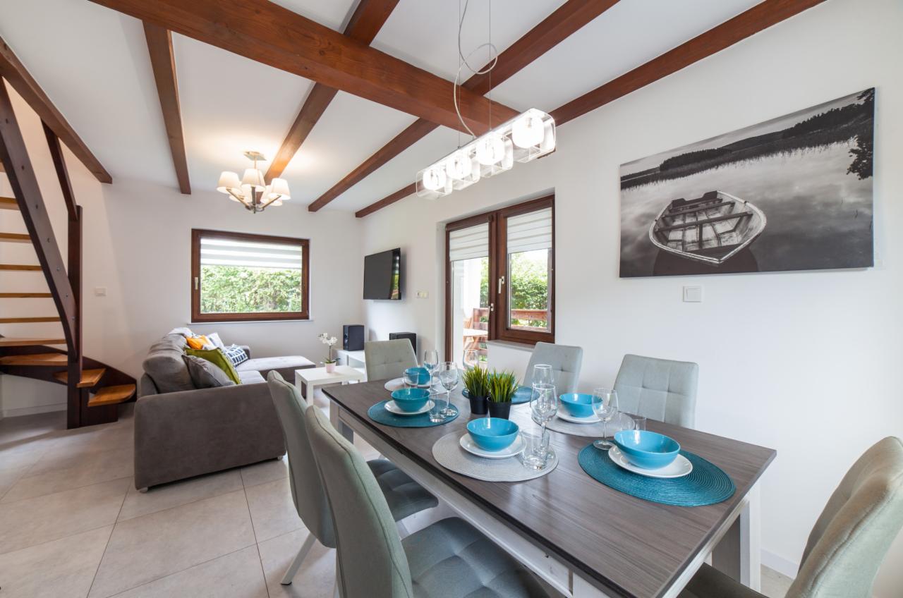 Jasne wnętrze jadalni z drewnianymi belkami stropowymi, stołem nakrytym turkusową zastawą i szarą sofą w tle.