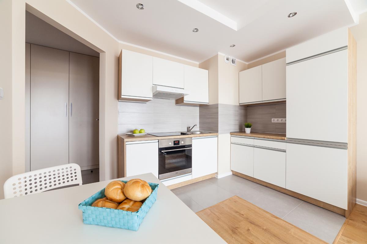 Jasna kuchnia z białymi szafkami i drewnianymi blatami, na stole błękitny koszyk z bułkami, widok na płytę indukcyjną i piekarnik.