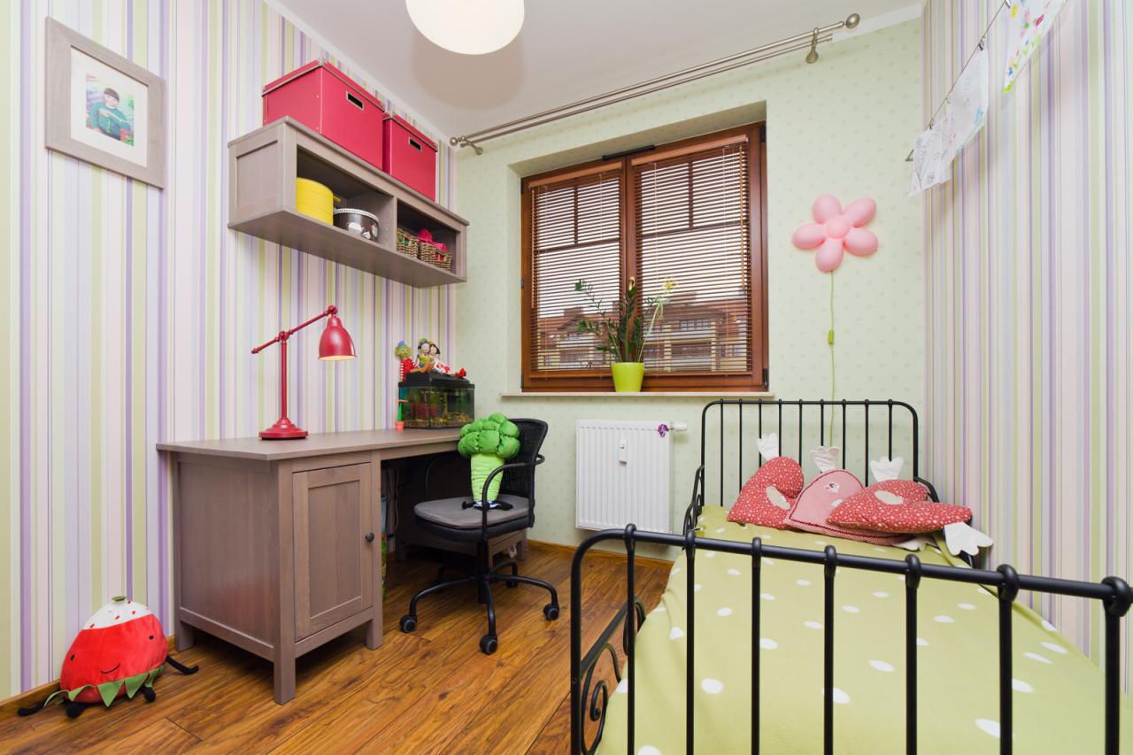 Pokój dziecięcy z biurkiem, łóżkiem z metalową ramą i akcentami w kolorze czerwonym, w tym lampka na biurku i pudełka na półce, akwarium z rybkami na biurku, ściany w paski i kropki.