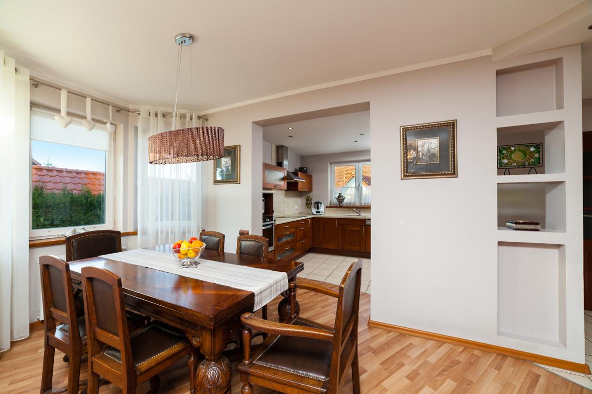 Jasne wnętrze jadalni z drewnianym stołem, krzesłami i owocami w misie, widok na kuchnię w tle, dekoracyjne półki w ścianie i okno z widokiem na czerwoną dachówkę.