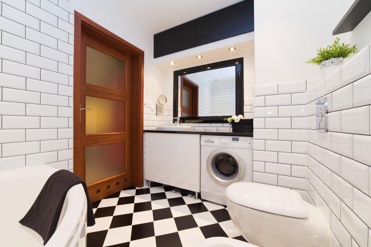 Biała łazienka z czarno-białą szachownicą na podłodze, białe kafelki typu metro na ścianach, drewniane drzwi z przeszkleniami i pralka obok umywalki.