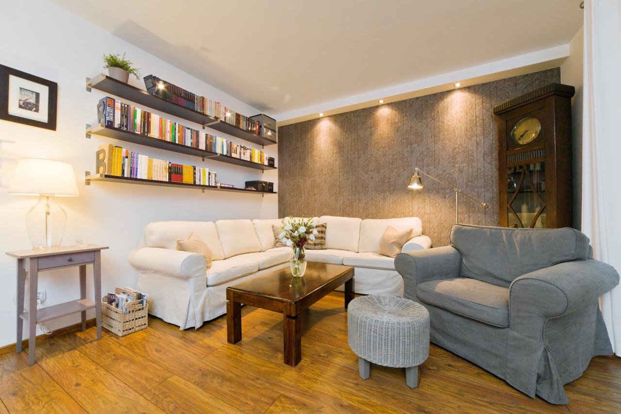 Jasny salon z narożną sofą, drewnianym stolikiem kawowym, zegarem stojącym i półkami z książkami, oświetlony punktowymi lampami.