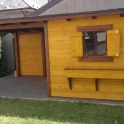 Żółta, drewniana altana z zadaszeniem na podwórku, z oknem z okiennicami i skrzynką na kwiaty, wykończona brązowymi elementami.