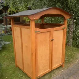 Drewniana, zamykana szafka ogrodowa z dwuspadowym daszkiem, umieszczona na trawniku obok chodnika.