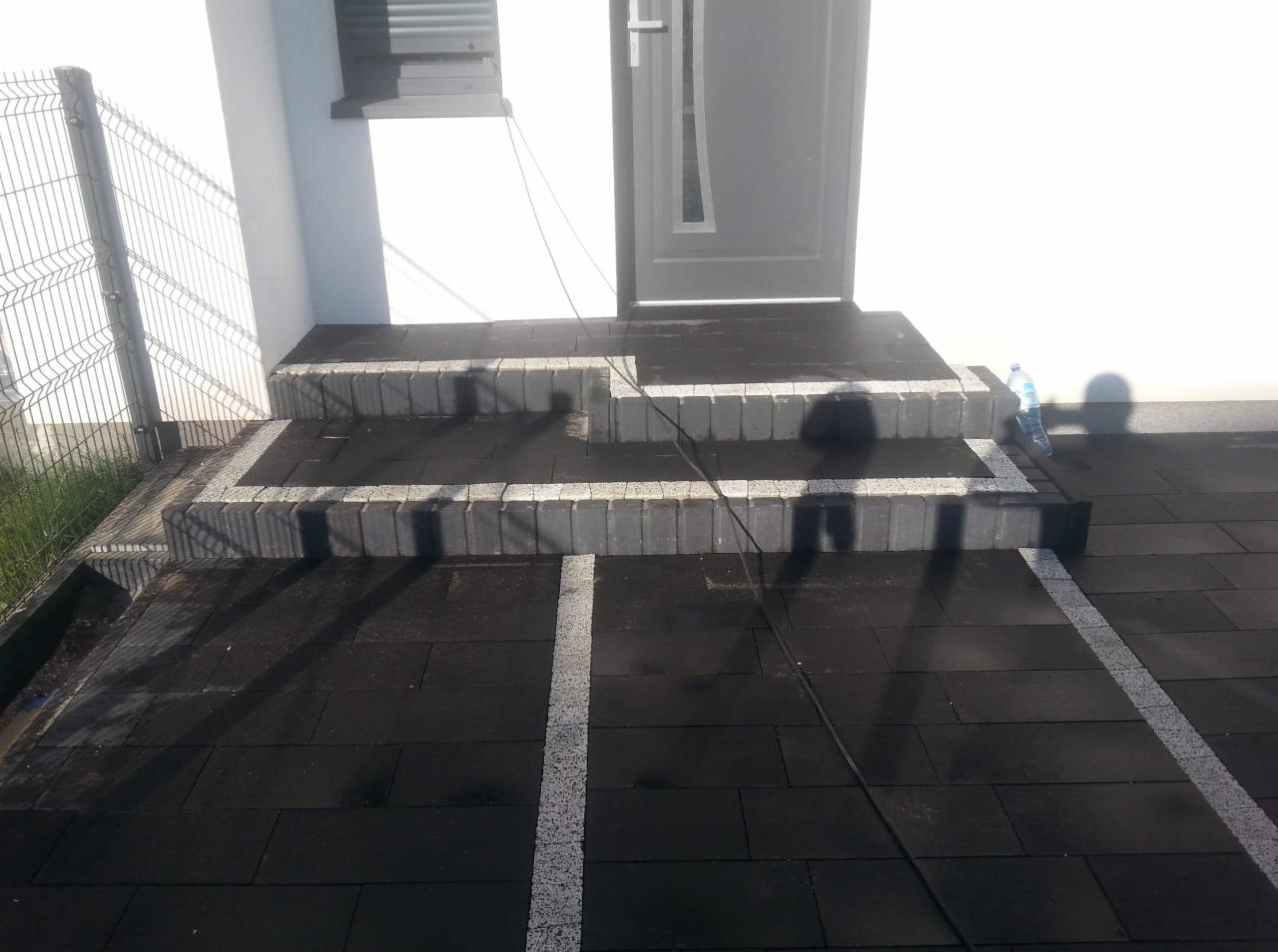 Schody wejściowe do domu wyłożone ciemną kostką brukową z jasnymi obrzeżami, widoczna butelka wody i cień osoby.