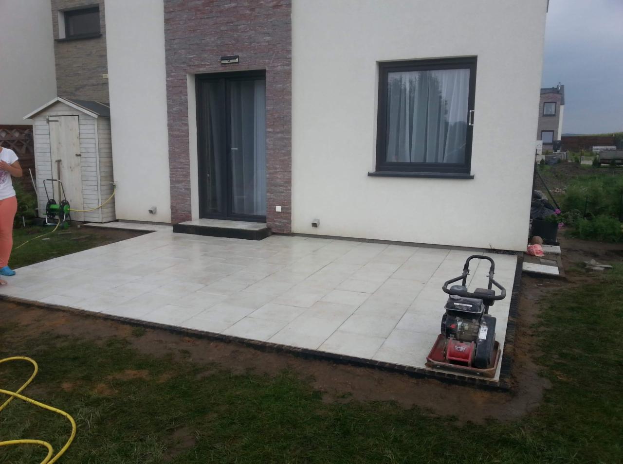 Jasne, kwadratowe płyty ułożone na tarasie przydomowym, z widoczną zagęszczarką w prawym dolnym rogu i fragmentem osoby po lewej stronie