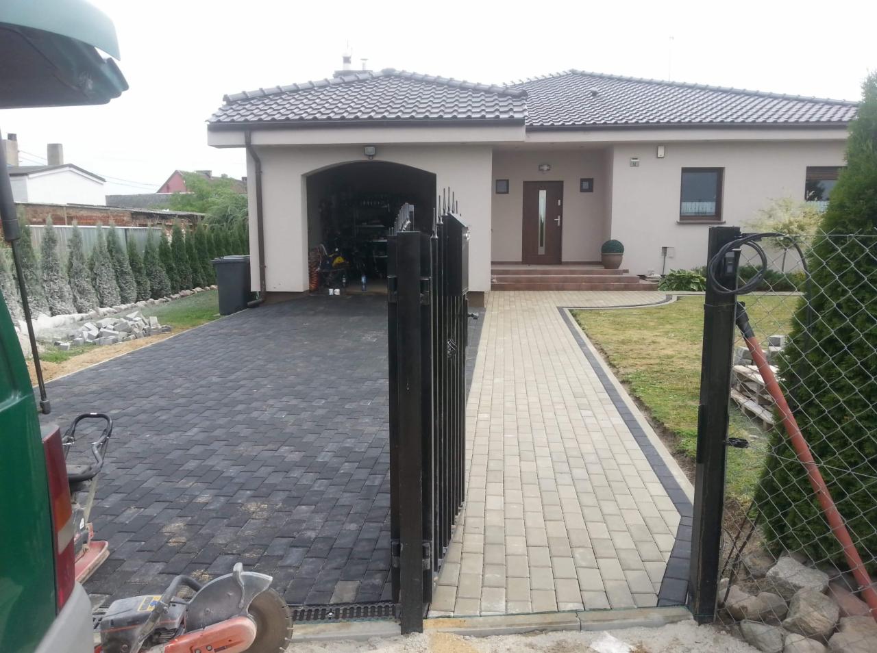 Ułożona kostka brukowa w dwóch odcieniach szarości prowadząca do domu z garażem, widoczna brama i narzędzia brukarskie na pierwszym planie.