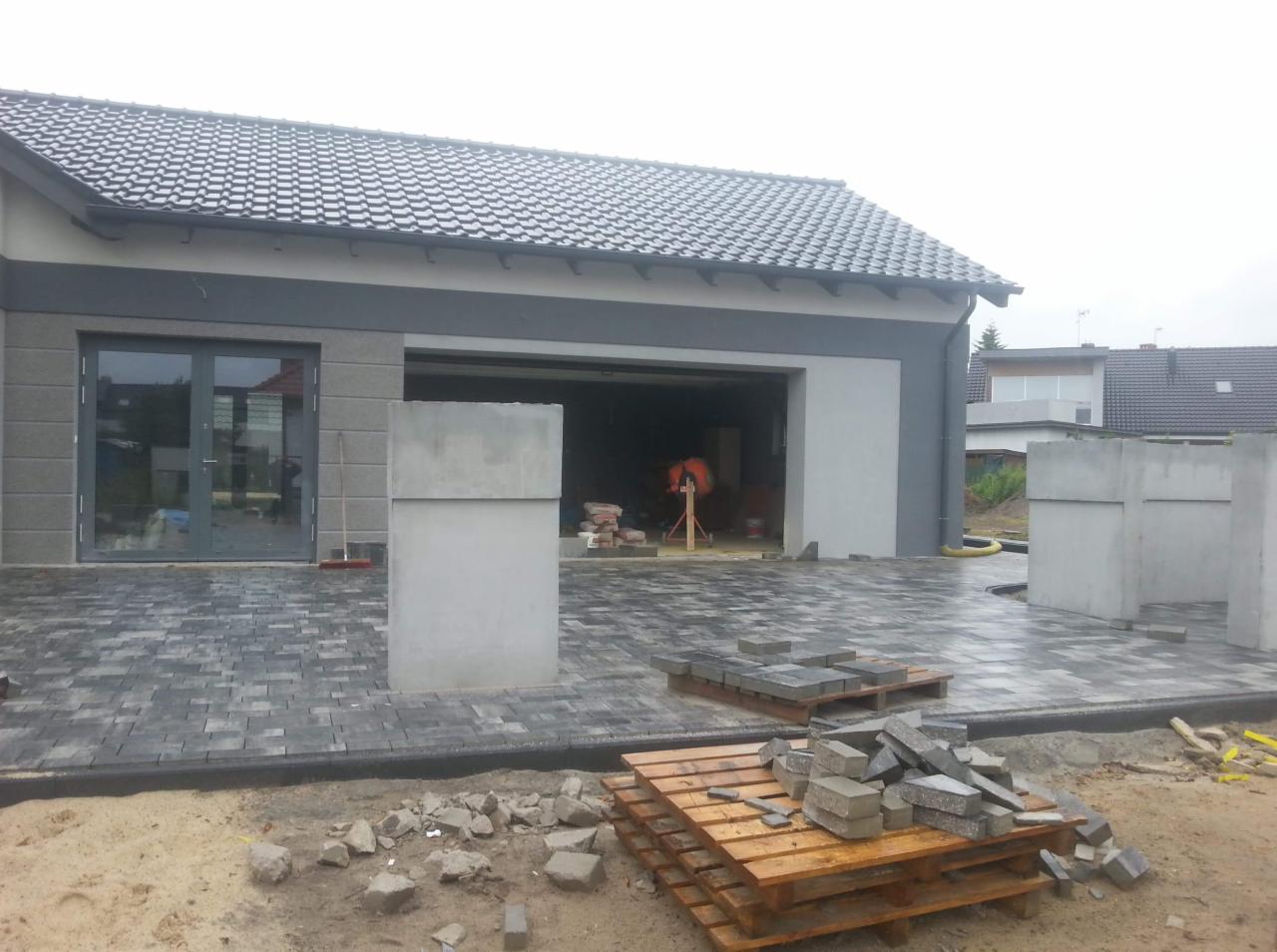 Układanie szarej kostki brukowej przed nowoczesnym domem z garażem; widoczne palety z kostką, betonowe słupki i betoniarka w garażu.