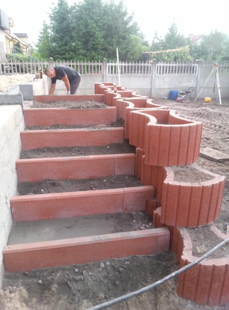 Budowa schodów zewnętrznych z czerwonej kostki, z ozdobnymi obrzeżami w kształcie fal, na podwórku z widocznym pracownikiem wyrównującym ziemię na stopniach.