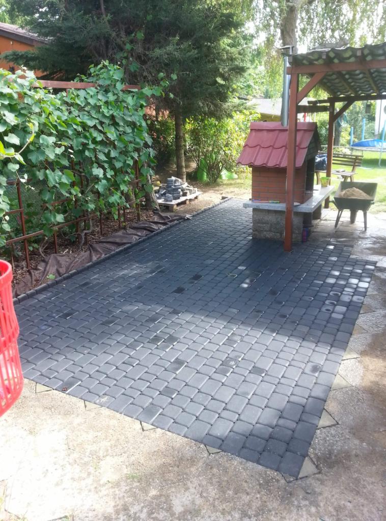 Nowo ułożona, ciemnoszara kostka brukowa na posesji, widoczny grill z czerwonym dachem, fragment winorośli pnącej się po konstrukcji oraz taczka z piaskiem w tle.