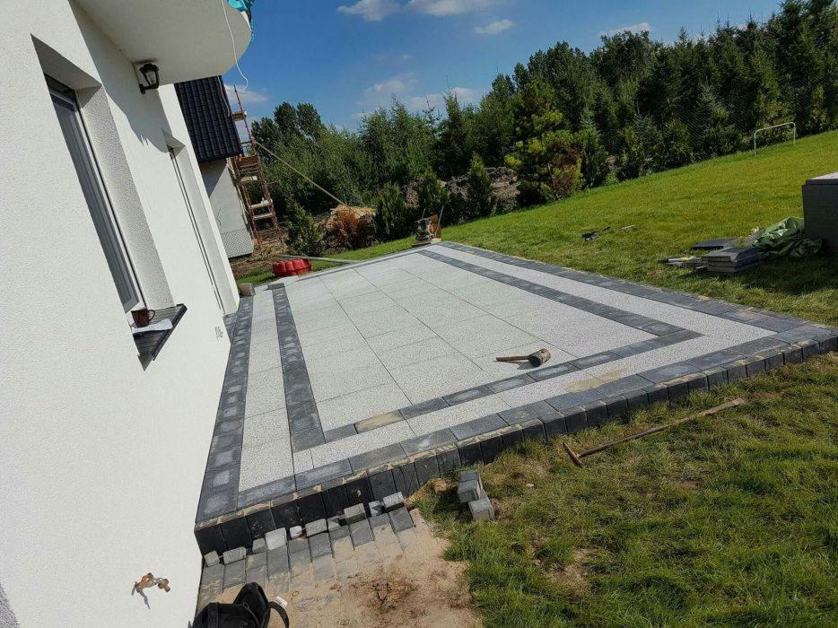 Wykończony taras z jasnoszarej kostki brukowej z ciemnoszarą obwódką, obok domu z trawnikiem i drzewami w tle, widoczny młotek brukarski.