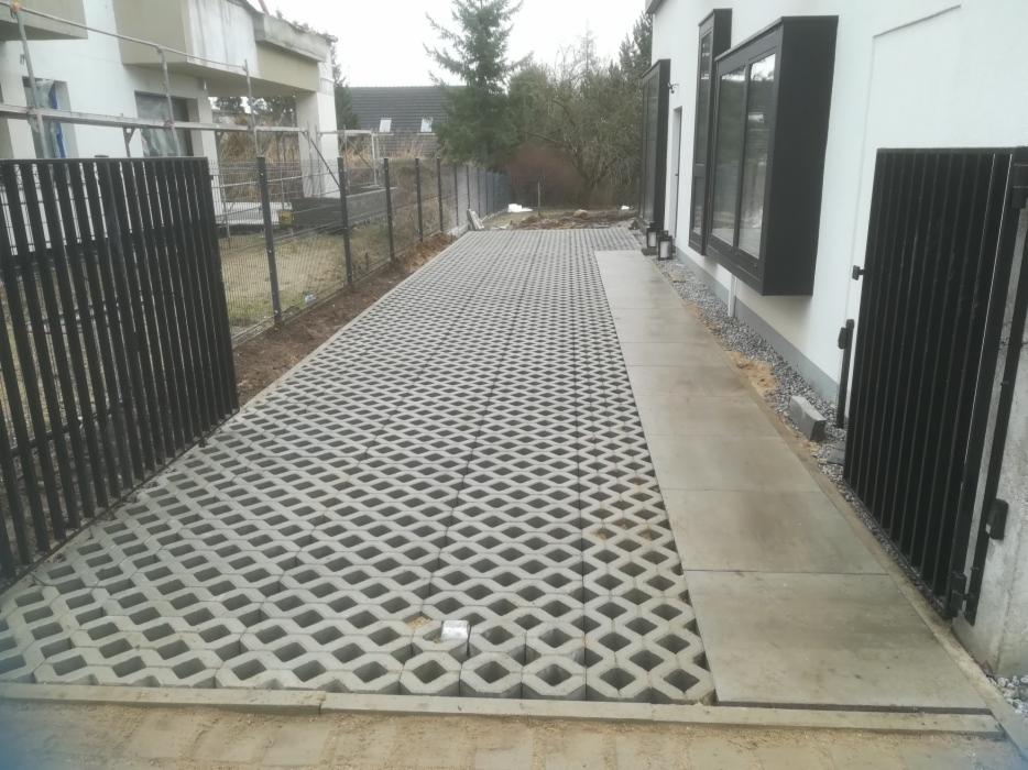 Ułożona szara kostka ażurowa na podjeździe, widoczna furtka i fragment domu z czarnymi oknami.