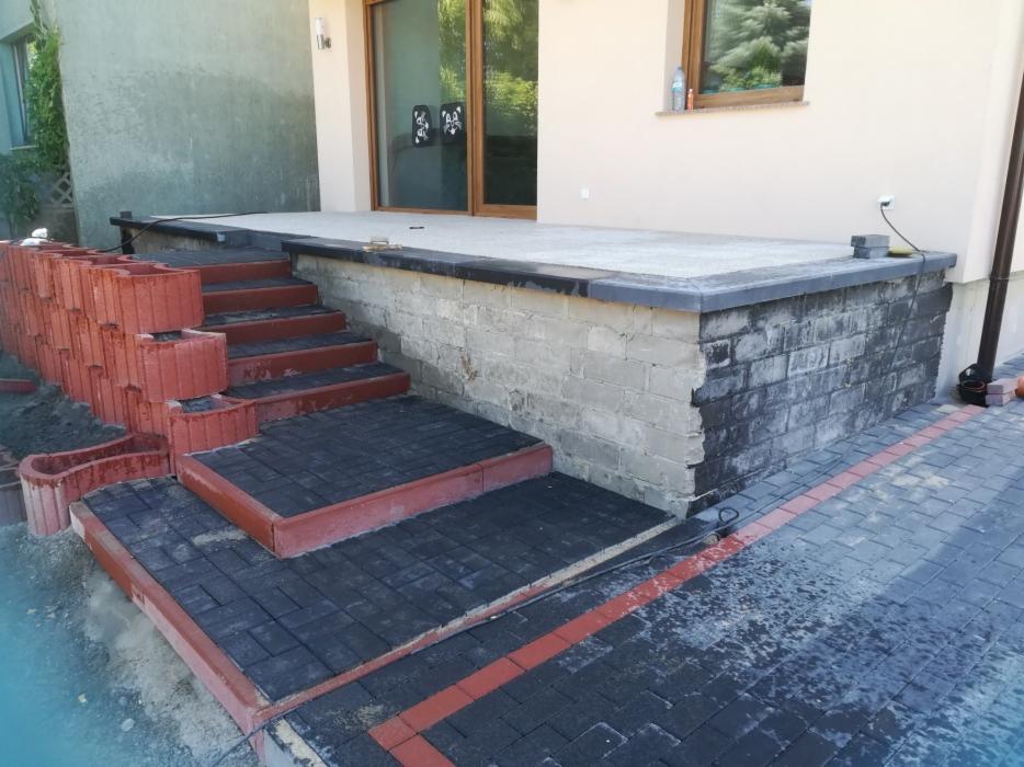 Budowa schodów wejściowych z kostki brukowej w kolorze grafitowym z czerwonym obramowaniem, widoczne donice z czerwonej kostki i fragment elewacji budynku.