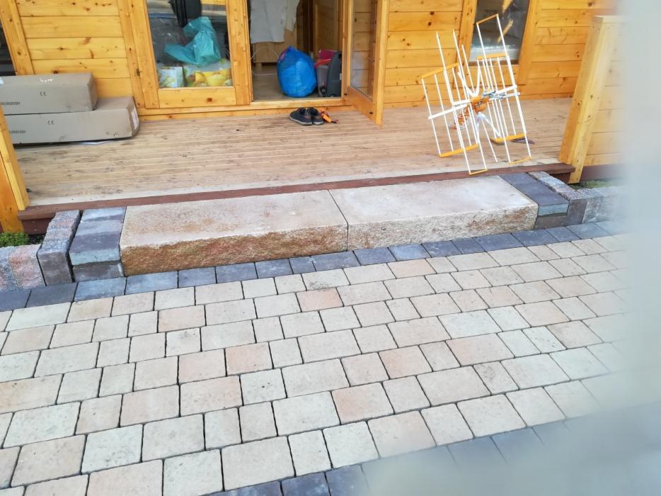 Ułożona kostka brukowa w odcieniach beżu i szarości prowadząca do drewnianego domku z tarasem, z kamiennymi stopniami wejściowymi.