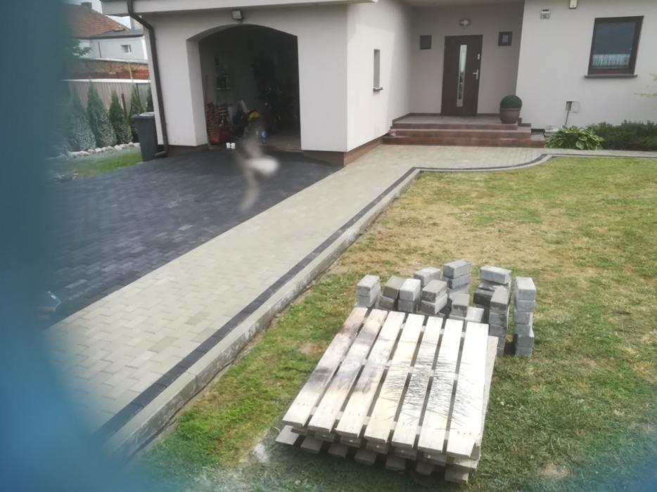 Ułożona ścieżka z jasnej kostki brukowej z ciemnym obramowaniem prowadząca do domu z podjazdem z ciemnej kostki, paleta z kostką obok trawnika.