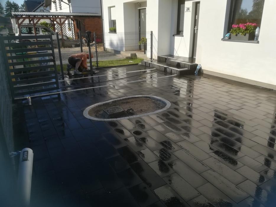 Układanie ciemnej kostki brukowej na posesji przed domem; widoczny pracownik w trakcie pracy, przygotowane miejsce na rabatę, odbicia w mokrej nawierzchni.