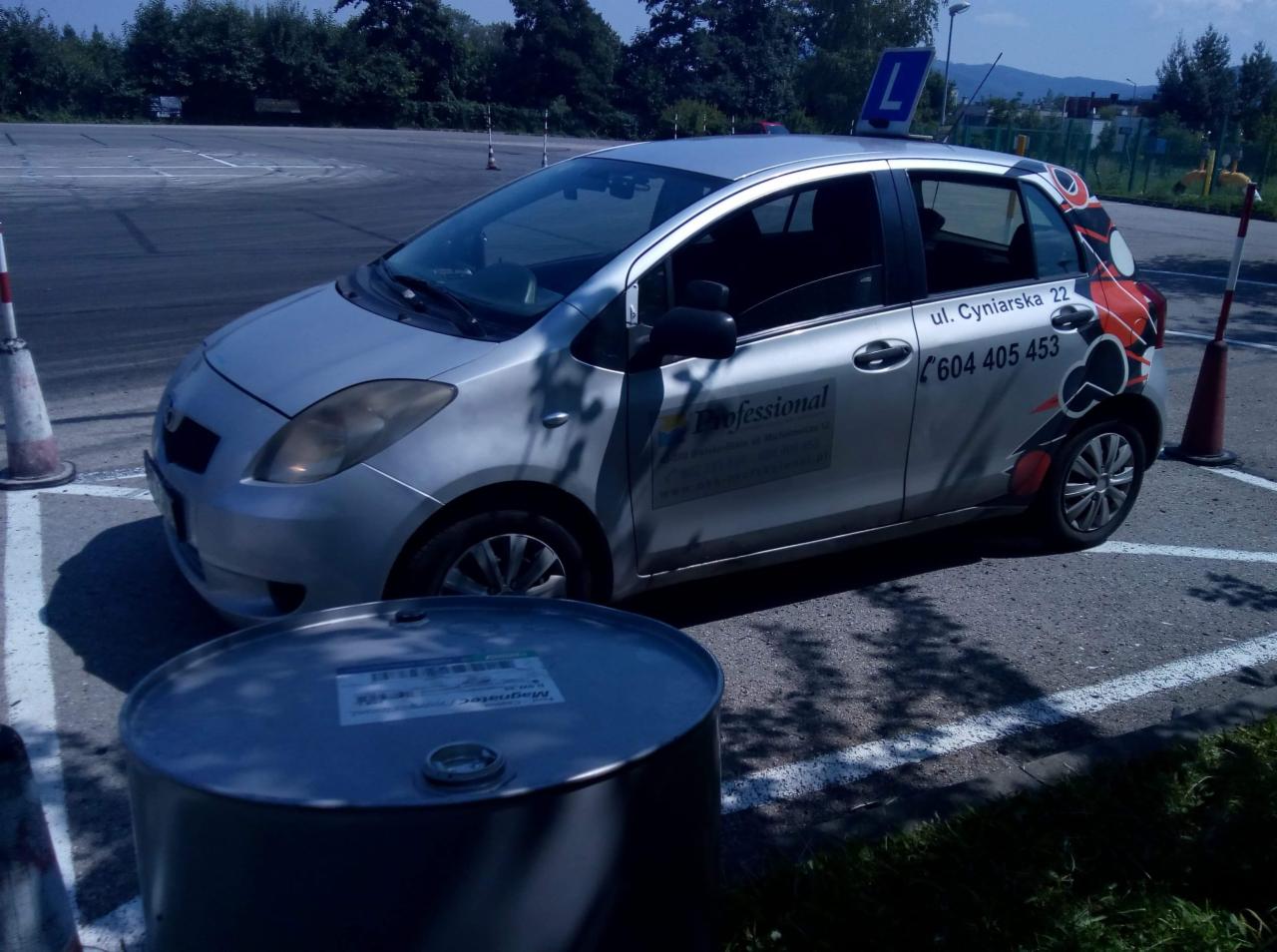 Srebrny samochód szkoły jazdy z naklejkami reklamowymi, stojący na placu manewrowym obok beczki i pachołków.