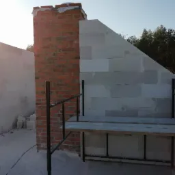 Wznoszenie muru domu z bloczków i komina z cegły, widoczne rusztowanie i fragment lasu w tle.