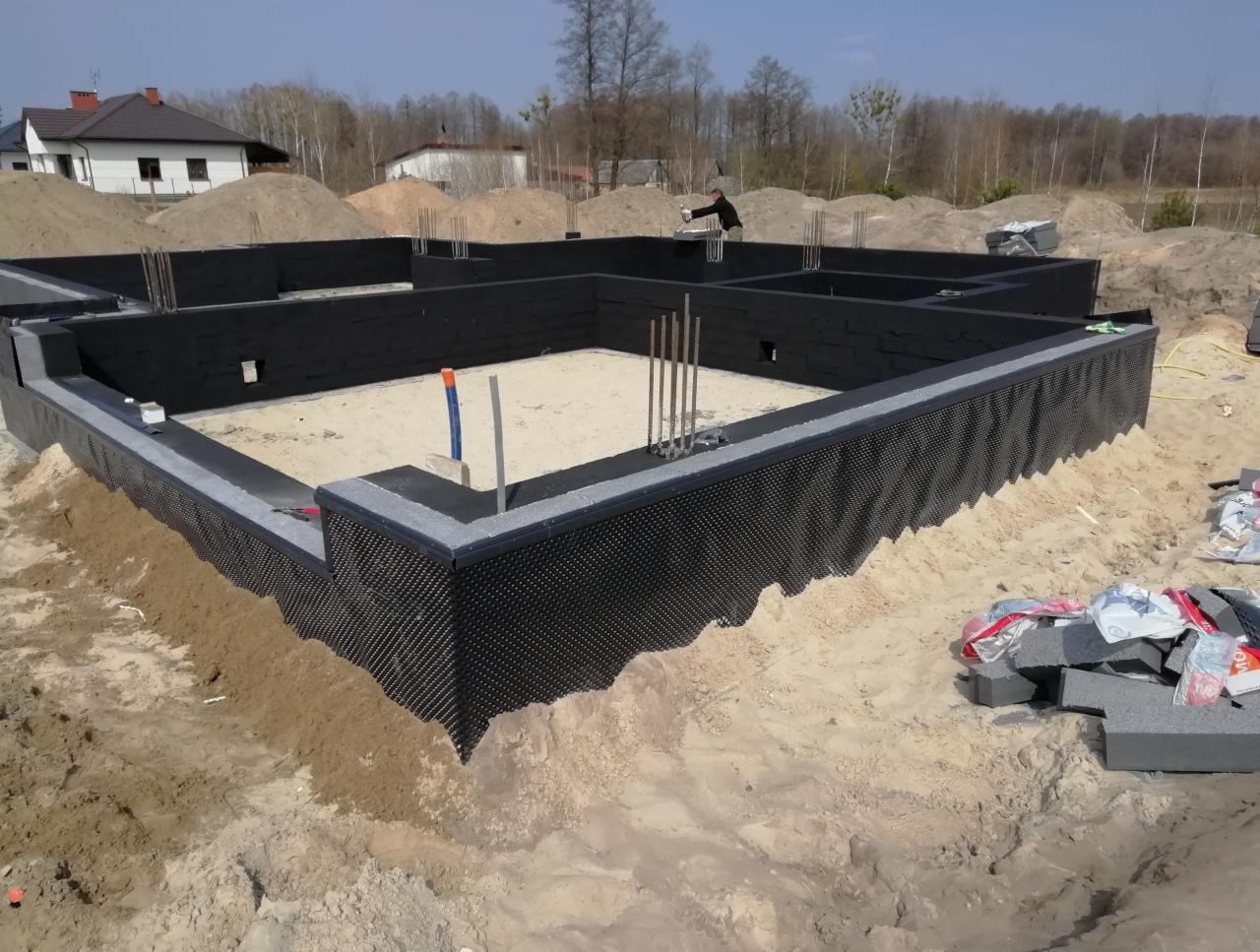 Zabezpieczone fundamenty domu jednorodzinnego z hydroizolacją, widoczne wystające pręty zbrojeniowe i częściowo zasypany wykop.