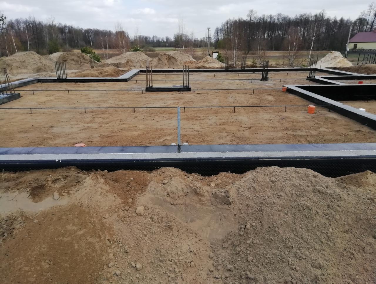 Zarys fundamentów domu jednorodzinnego z wystającymi prętami zbrojeniowymi, widok z poziomu gruntu na przygotowany teren budowy.