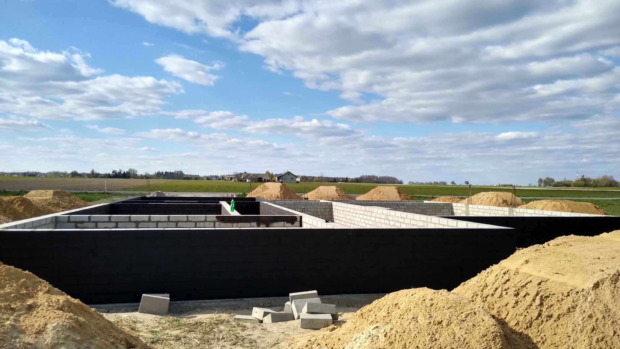 Wylane fundamenty z bloczków betonowych, zabezpieczone czarną izolacją, z widocznymi pryzmami piasku, w tle krajobraz z polami i zabudowaniami pod błękitnym, pochmurnym niebem.
