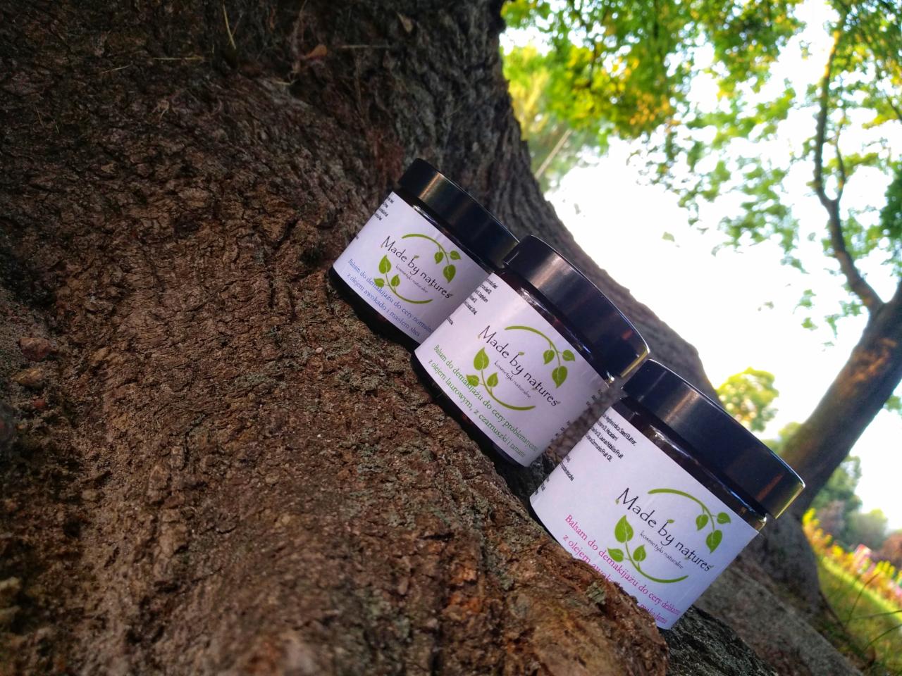 Trzy słoiczki balsamu do demakijażu 'Made by natures' z różnymi olejkami, ustawione na korze drzewa na zewnątrz.
