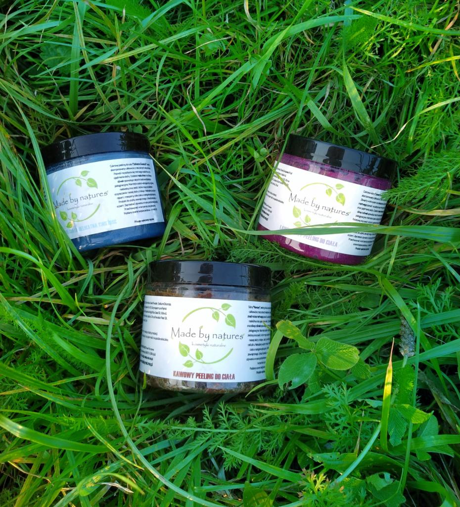 Trzy słoiczki z peelingami do ciała marki Made by Natures, w kolorach niebieskim, różowym i brązowym, ułożone na tle zielonej trawy. Słoiczek z brązowym peelingiem na pierwszym planie, z napisem...