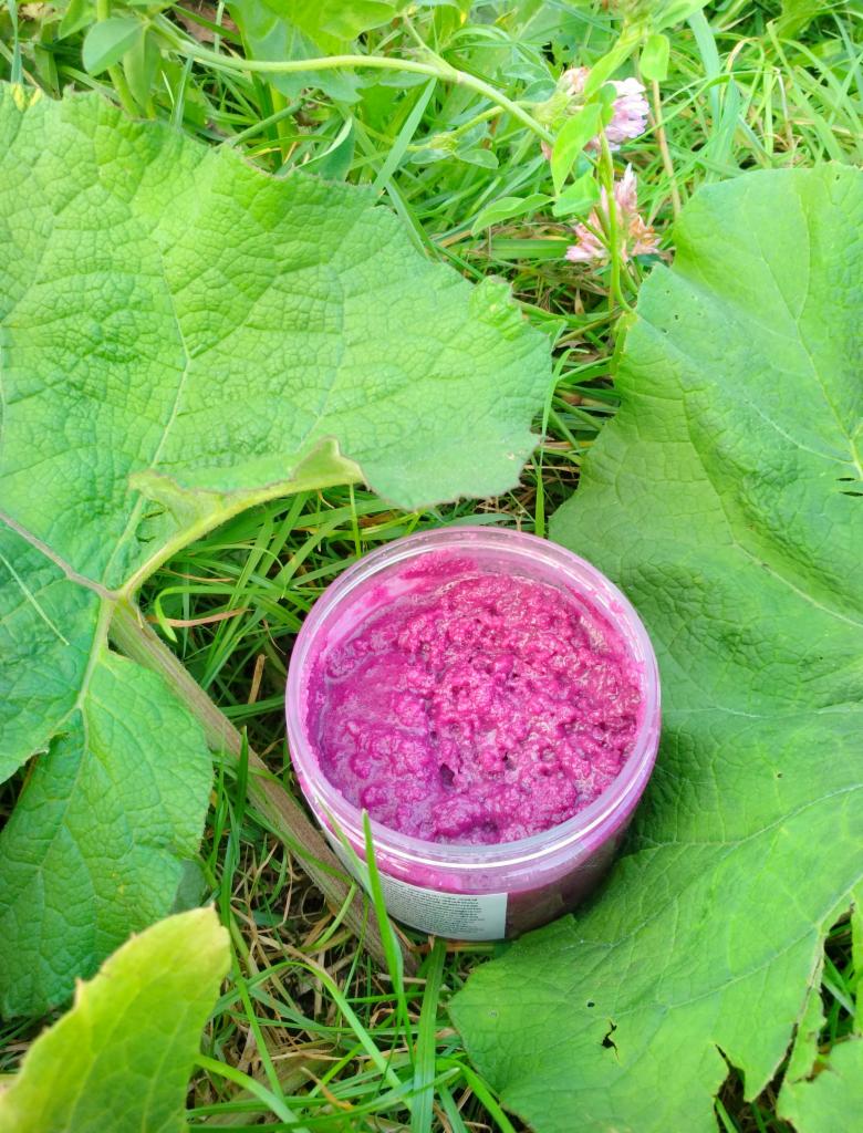 Różowy peeling w przezroczystym słoiczku, umieszczony na trawie obok dużych, zielonych liści.