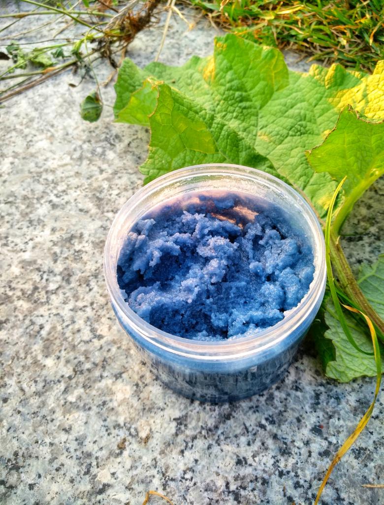Niebieski peeling cukrowy w plastikowym słoiczku na tle szarego kamienia i zielonych liści.