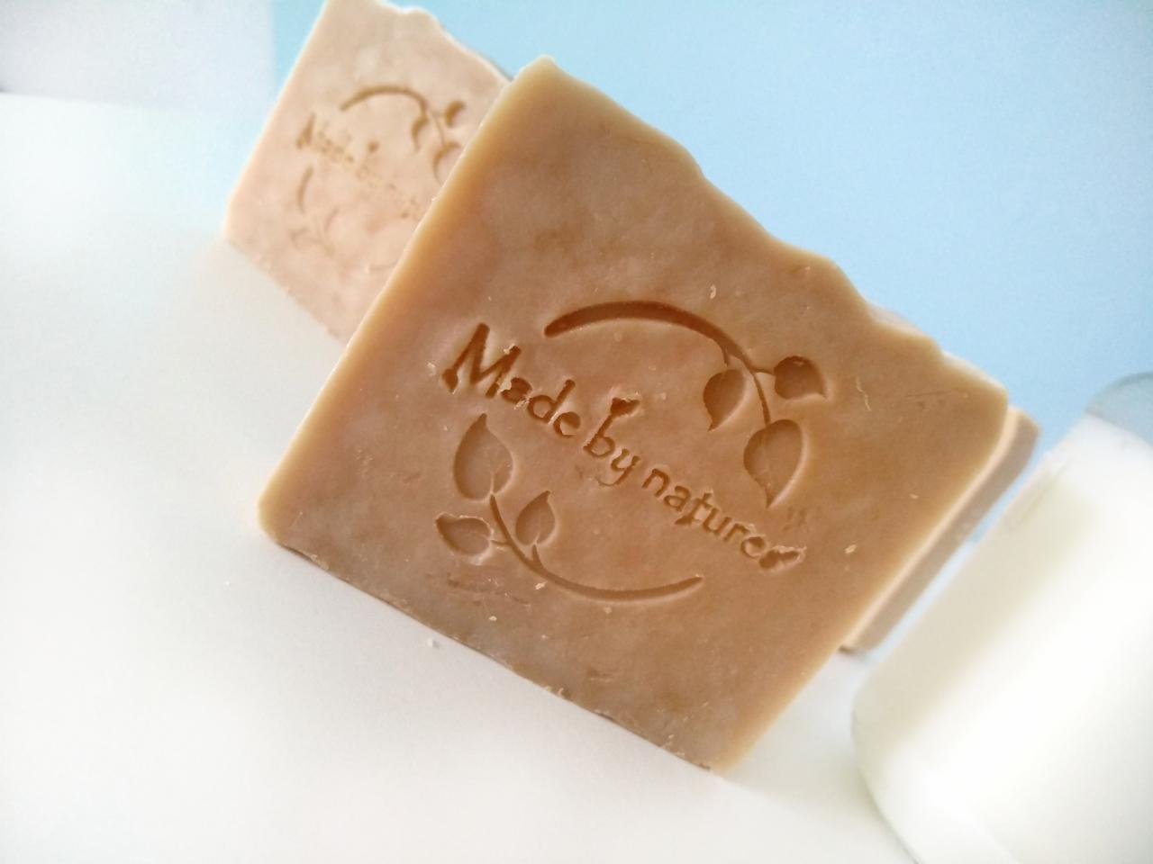 Dwa kwadratowe mydła z wytłoczonym napisem 'Made by nature' i motywem liści, obok szklanki z białym płynem na białym tle z błękitnym akcentem.