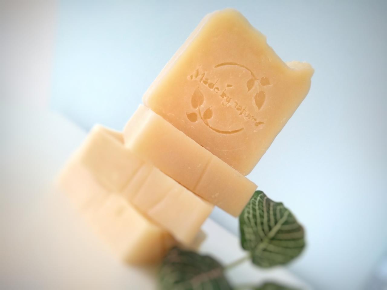 Stos kostek naturalnego, beżowego mydła z wytłoczonym napisem 'Made by nature' i motywem liści, ułożonych na białym tle z zielonym liściem rośliny.