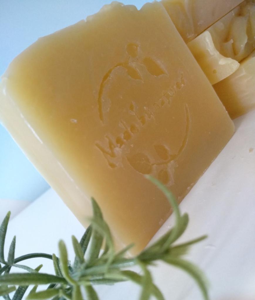 Kwadratowa kostka naturalnego mydła z odciśniętym logo firmy, ułożona obok gałązki rozmarynu, na białym tle.