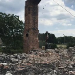 Ceglany komin częściowo zburzony, z wiszącą górną sekcją, gruzowisko u podstawy, w tle koparka z młotem pneumatycznym i zieleń.