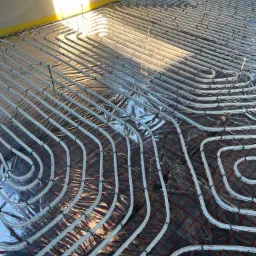 Białe rury ogrzewania podłogowego ułożone w pętle na folii z siatką, widoczne mocowania i fragment ściany z oknem w tle.