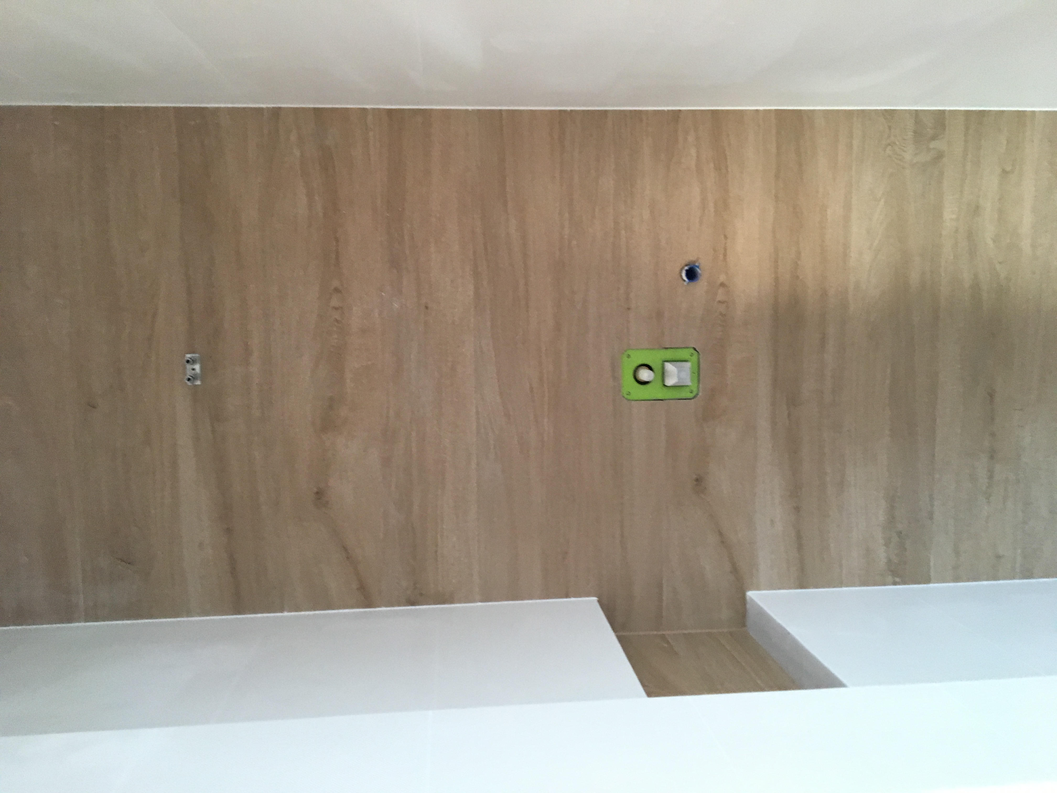 Ściana wyłożona płytkami imitującymi drewno w trakcie montażu, widoczne puszki elektryczne i otwór na rurę, biały sufit i blat poniżej.