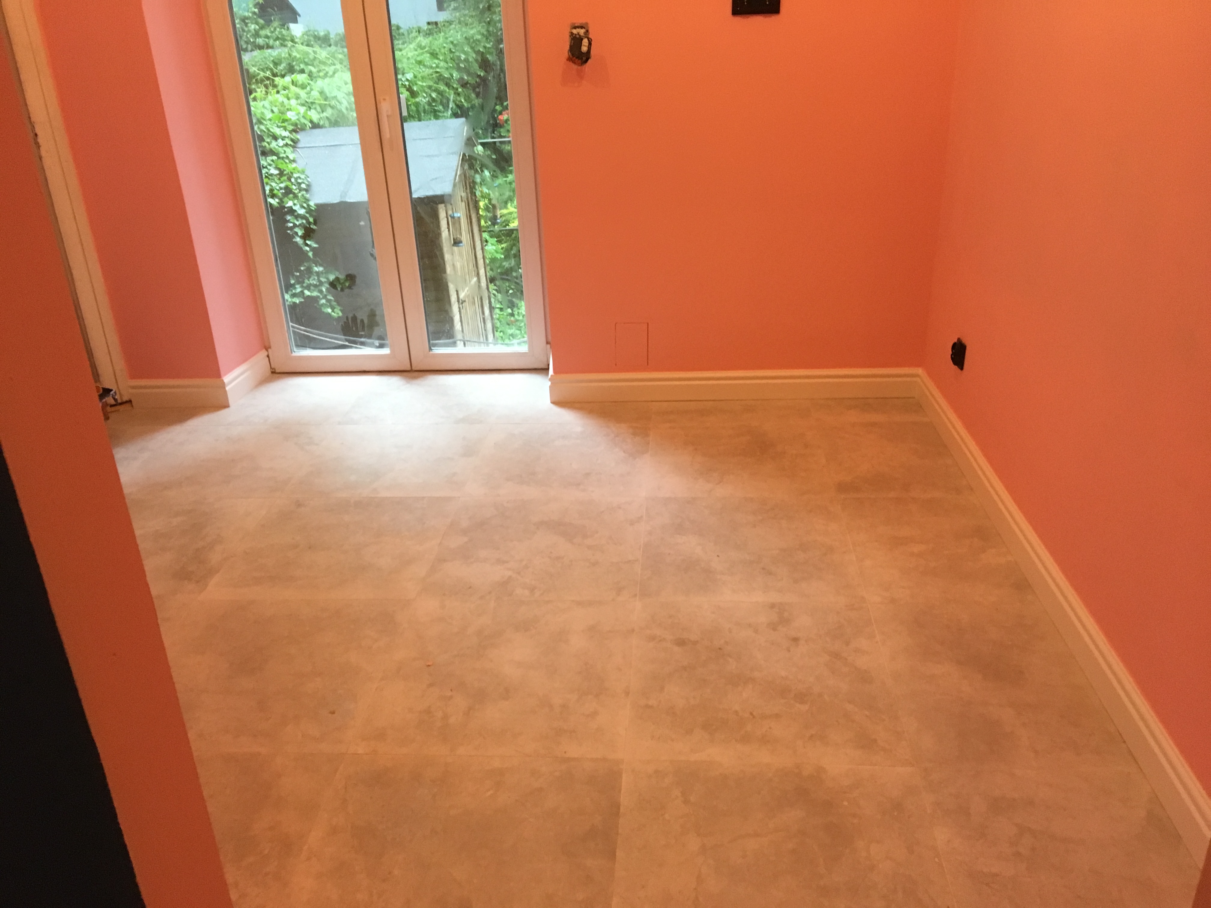 Wykończone wnętrze z pomarańczowymi ścianami i jasnobeżową podłogą z płytek, widok na zewnątrz przez dwuskrzydłowe okno balkonowe.