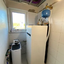 Biała pompa ciepła z niebieskim zbiornikiem wyrównawczym i manometrem, zainstalowana w pomieszczeniu wyłożonym białymi płytkami, widoczne rury i instalacja wodna, sufit z widocznymi fioletowymi...