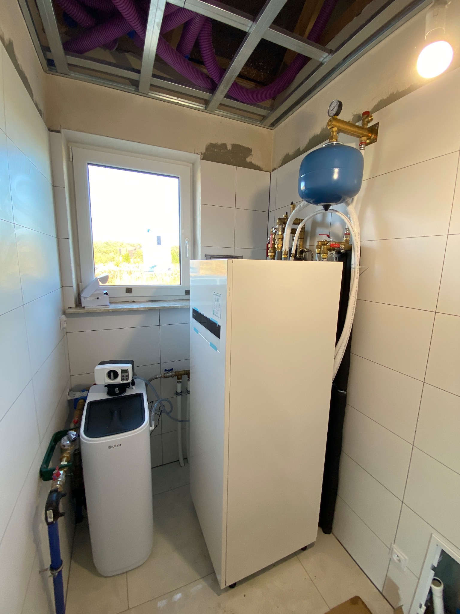 Biała pompa ciepła z niebieskim zbiornikiem wyrównawczym i manometrem, zainstalowana w pomieszczeniu wyłożonym białymi płytkami, widoczne rury i instalacja wodna, sufit z widocznymi fioletowymi...