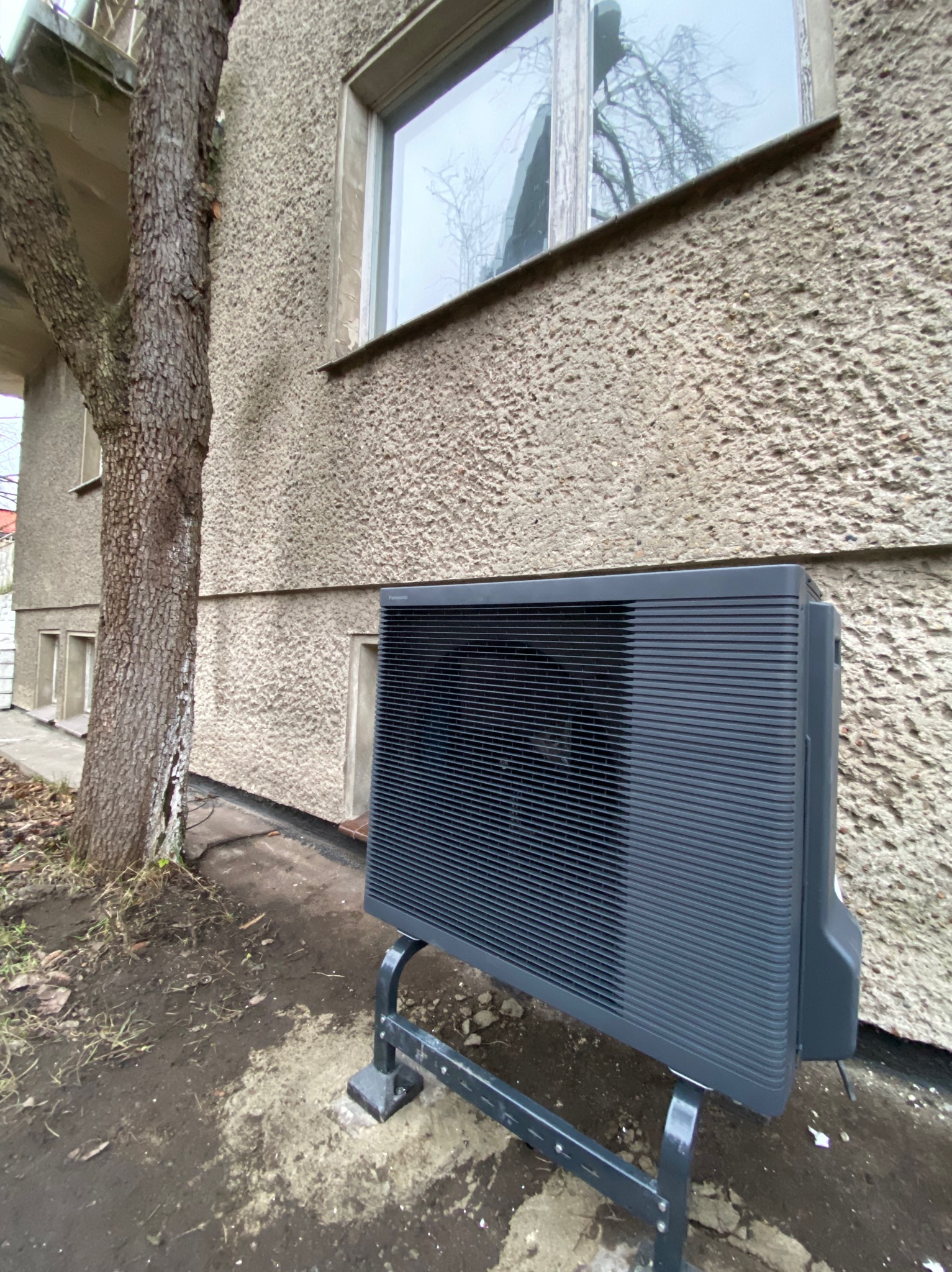 Zewnętrzna jednostka pompy ciepła Panasonic Aquarea zamontowana na metalowym stelażu przy ścianie budynku z oknem i obok drzewa.