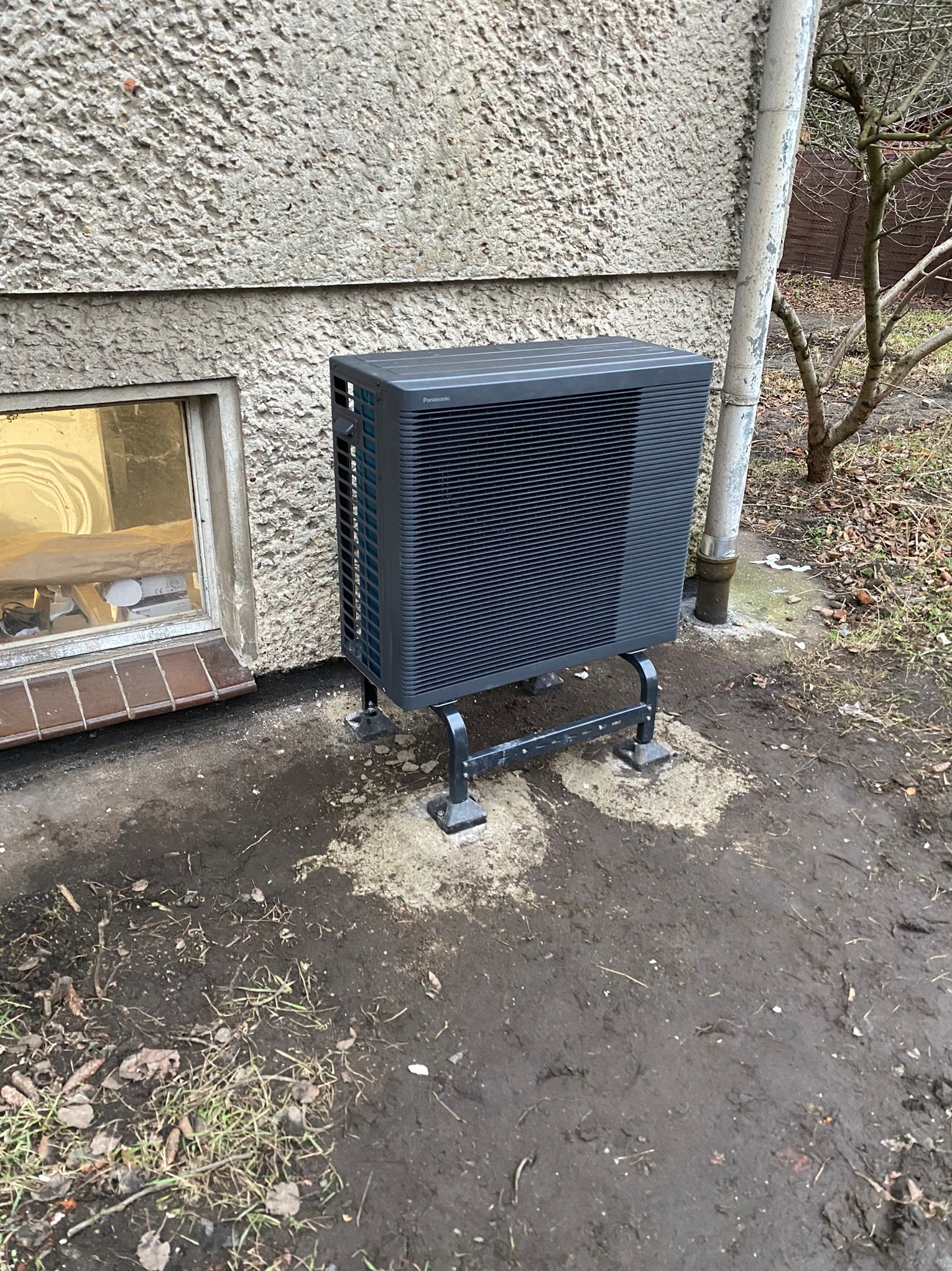 Zewnętrzna jednostka klimatyzacji Panasonic zamontowana na metalowym wsporniku przy ścianie budynku z oknem i rurą spustową, widoczne zabrudzenia na ziemi.