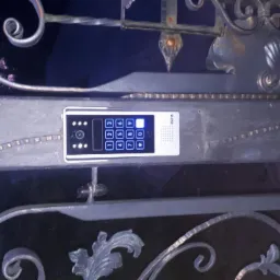 Zewnętrzny panel wideodomofonu z klawiaturą numeryczną, zamontowany na metalowej, ozdobnej bramie posesji, widoczne elementy dekoracyjne bramy.