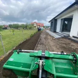Widok z perspektywy operatora na glebogryzarkę przygotowującą pas ziemi wzdłuż ogrodzenia, z widocznym fragmentem nowego domu i pracującymi osobami w tle.