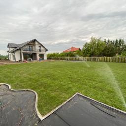 M-Lan - Rozległy trawnik nawadniany przez automatyczny system zraszający, widoczny dom z nowoczesną architekturą w tle, oraz fragment przygotowanej rabaty z obrzeżem i rozłożoną agrowłókniną.