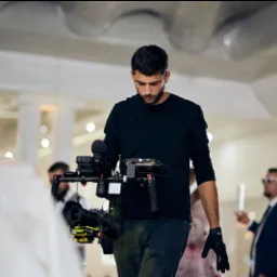 Młody mężczyzna w czarnym ubraniu z profesjonalnym sprzętem do nagrywania wideo na stabilizatorze, patrzący w dół, w tle widoczne osoby na uroczystości.