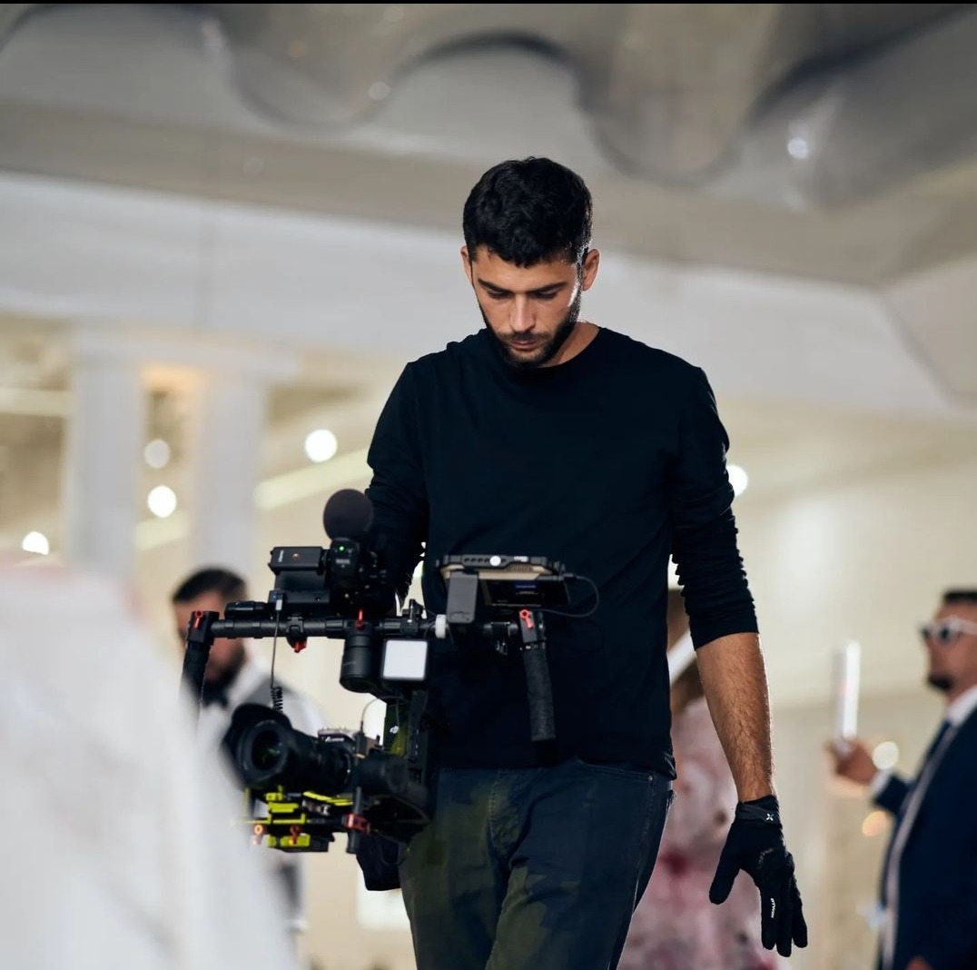 Młody mężczyzna w czarnym ubraniu z profesjonalnym sprzętem do nagrywania wideo na stabilizatorze, patrzący w dół, w tle widoczne osoby na uroczystości.