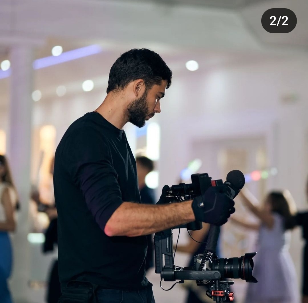 Mężczyzna z brodą w czarnej koszulce obsługuje profesjonalny stabilizator kamery z mikrofonem na tle sali z ludźmi.